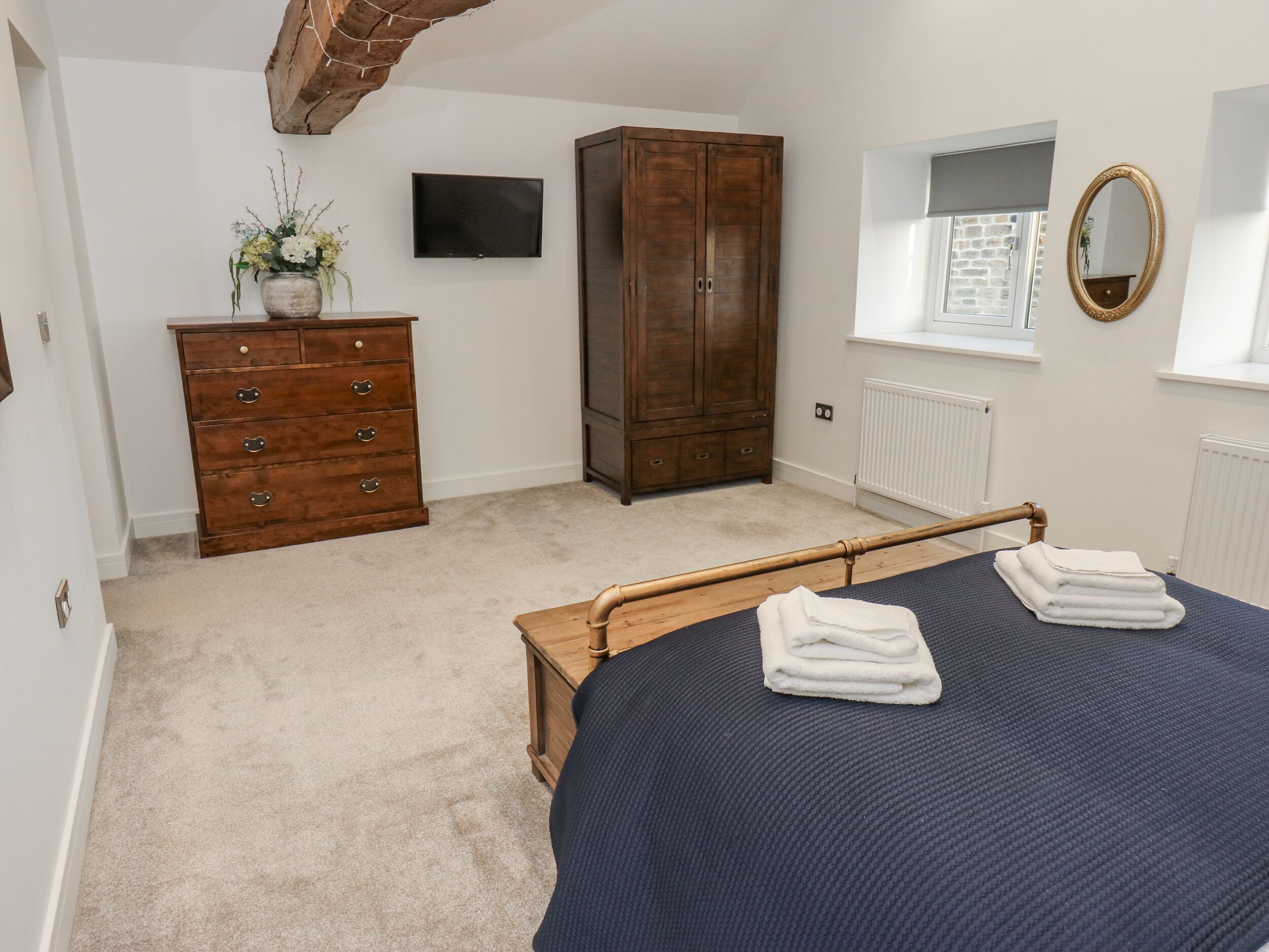 Een slaapkamer met een bed, een houten dressoir, een kledingkast, een televisie, een spiegel en een raam met uitzicht op een gebouw van baksteen.