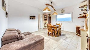 Ein Wohnzimmer mit Sofa, Esstisch und Stühlen, eine Küchenzeile mit Fenster sowie ein Deckenventilator.