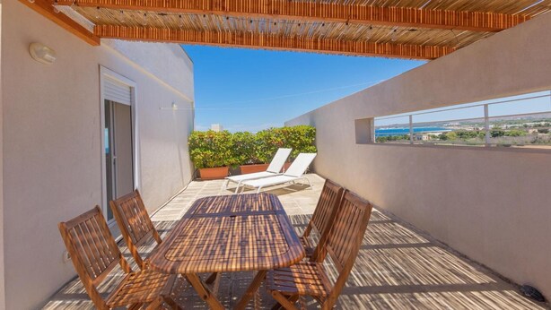 Eine überdachte Terrasse im Freien mit Tisch und Stühlen, Blick auf das Meer und ein Gebäude mit Tür und Fenstern.