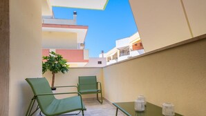 Een klein balkon met twee groene stoelen en een tafel, een bloempot, en een helder uitzicht op omliggende gebouwen.