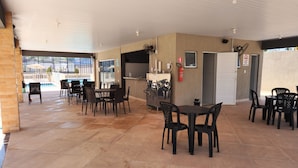 A covered outdoor area with tiled flooring, black plastic chairs, and tables. There is a pool visible through the glass enclosure in the background.