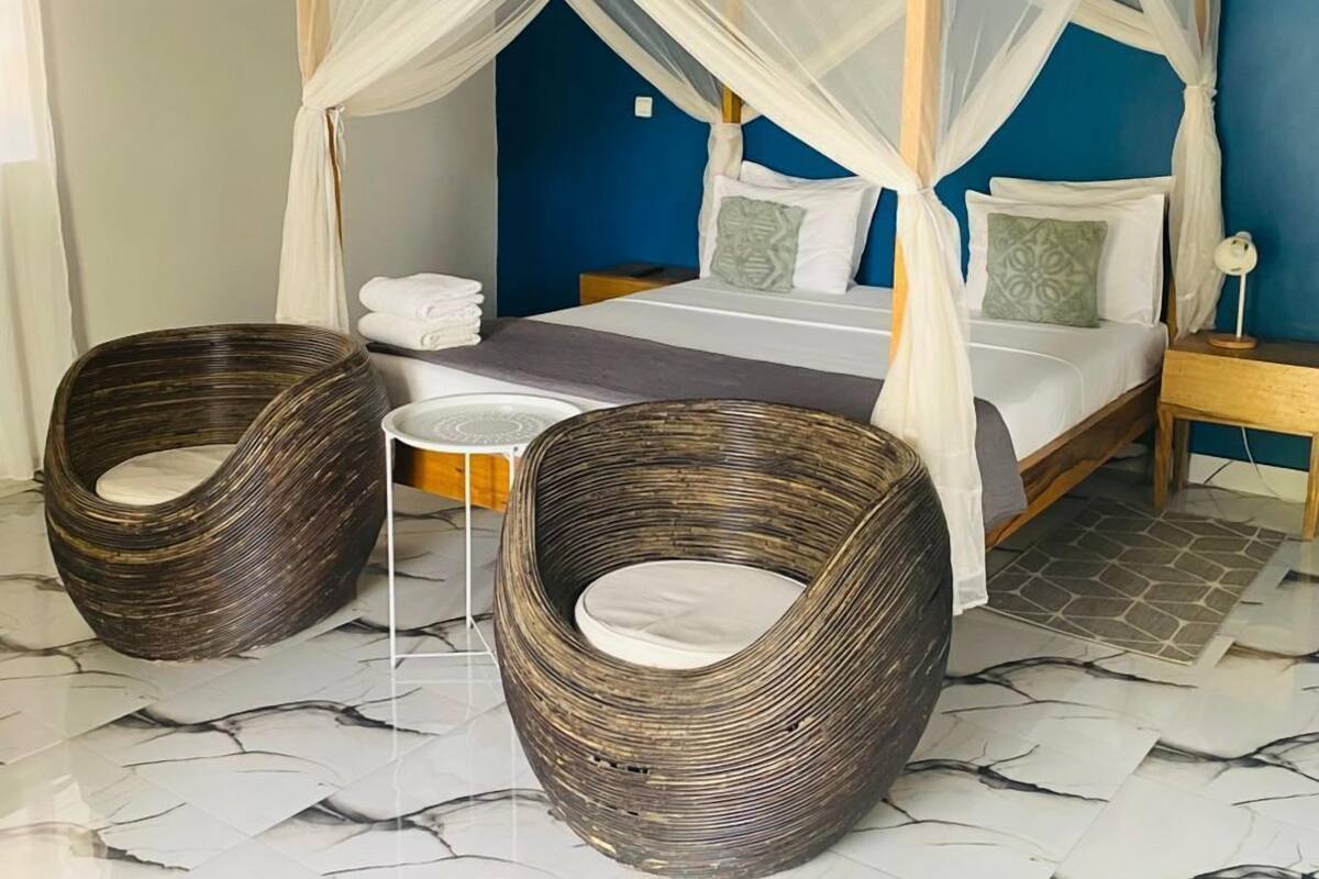 A four-poster bed with a canopy, two wicker chairs, a small table, and a nightstand with a lamp.