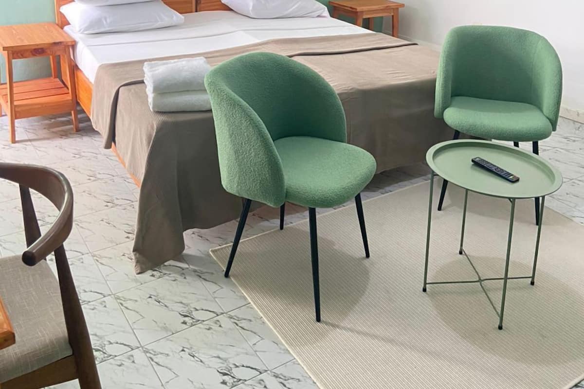 A room with a bed, two green chairs, a small table, a hanging light fixture, and an air conditioning unit.