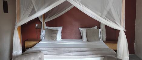A bed with a white canopy, two grey upholstered chairs, a small round wooden table, and a wall-mounted fan.