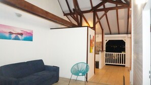 Ein Zimmer mit einem dunklen Sofa, einem türkisblauen Sessel, einem Sonnenuntergangsbild und einem Balkon mit Geländer.