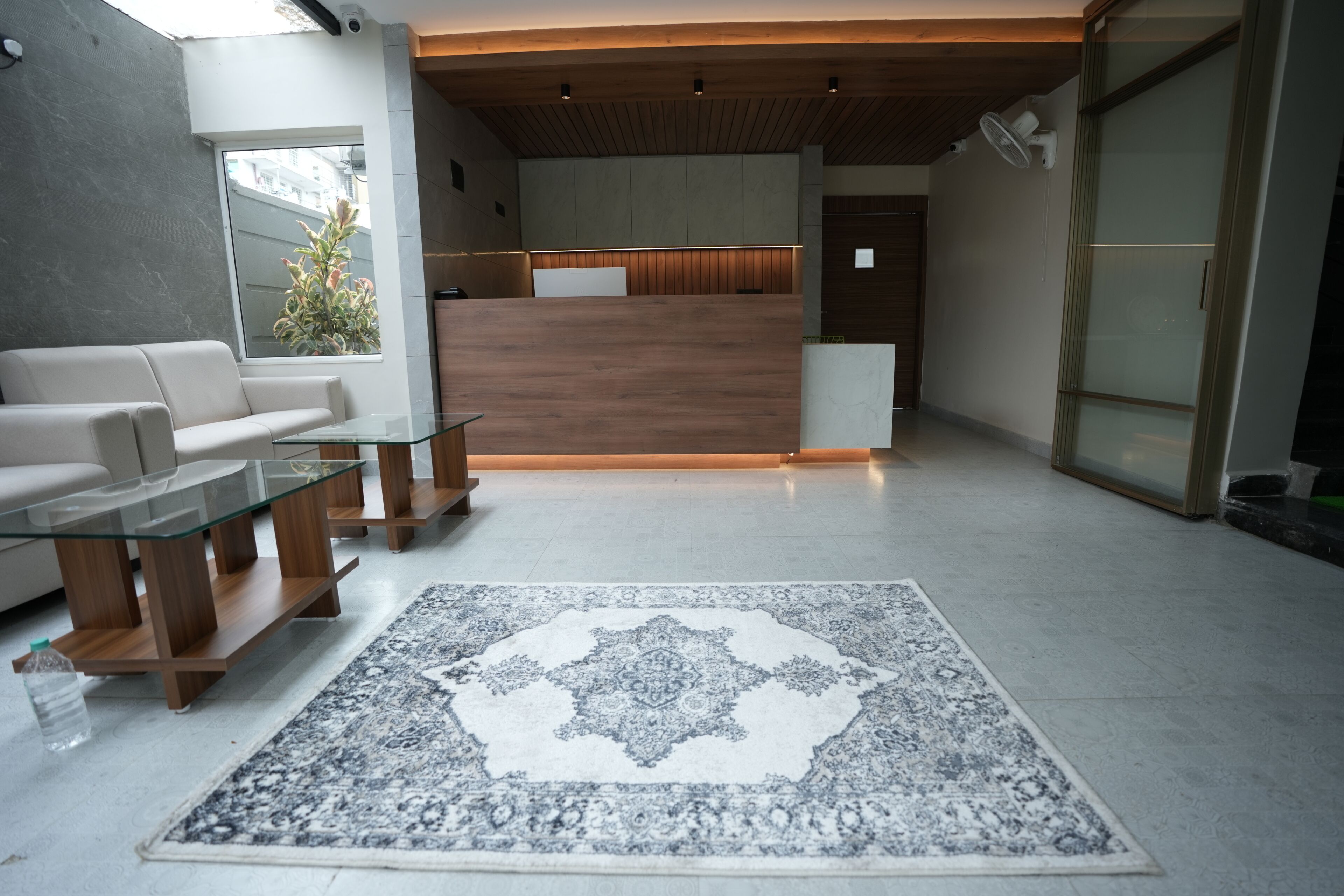 A modern reception area with a wooden reception desk, a glass coffee table, a patterned rug, and a sofa.