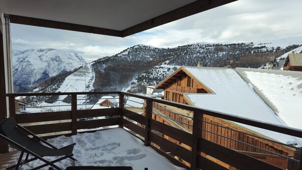 A wooden deck with a snow-covered ground, a wooden railing, and a view of a mountain landscape with houses. - Parenthèses A12 by Avecoeur (Huez)