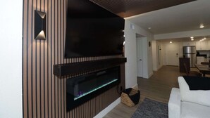 Ett modernt vardagsrum med en platt-TV som är monterad på en väggpanel av trä, en inbyggd öppen spis och ett kök i bakgrunden.