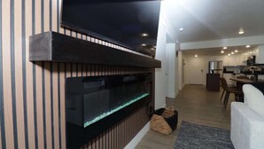 Ett modernt vardagsrum med en stor platt-TV som är monterad på en vägg av trä, ett inbyggt akvarium, en matplats med bord och stolar samt ett kök i bakgrunden.