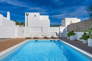 Piscina com cadeiras de banho brancas, um edifício branco com telhado plano e céu azul claro.