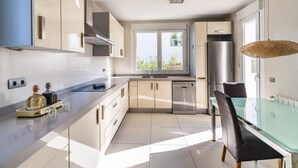 A modern kitchen with a dishwasher, a refrigerator, and a dining area with a glass table and chairs. - New Villa Noa 5 Bedrooms Private Ibiza Escape in Jesús (Santa Eulalia del Riu)