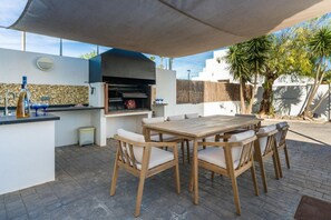 Uma cozinha externa com churrasqueira, mesa de jantar e cadeiras, cercada por um pátio com vista para um edifício branco e palmeiras.