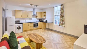 Une cuisine moderne avec des meubles en bois, un four à micro-ondes, un four et un lave-linge. Le salon est agrémenté d’un banc en bois et d’un tapis coloré.