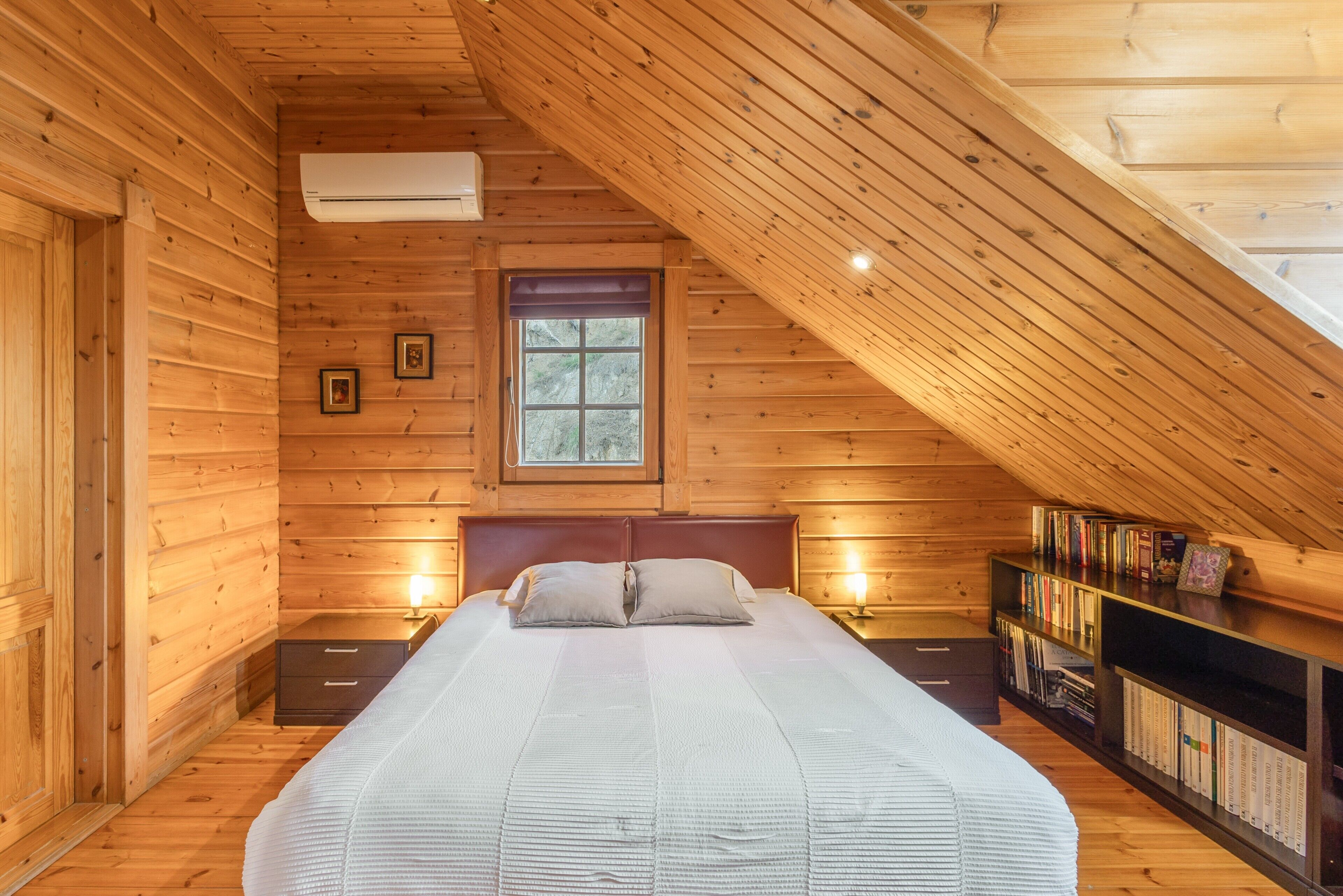 Un dormitorio de cabaña de madera con techo inclinado, una cama con almohadas, lámparas de noche, una ventana y una estantería.