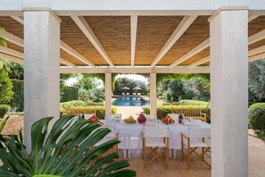 Een overdekt eetgedeelte met uitzicht op een zwembad, beschermd door een houten pergola, omringd door groen.