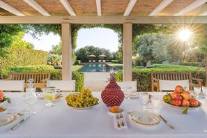 Un espace de restauration en plein air avec vue sur la piscine, des tables dressées avec de la vaisselle en verre et des présentoirs de fruits.