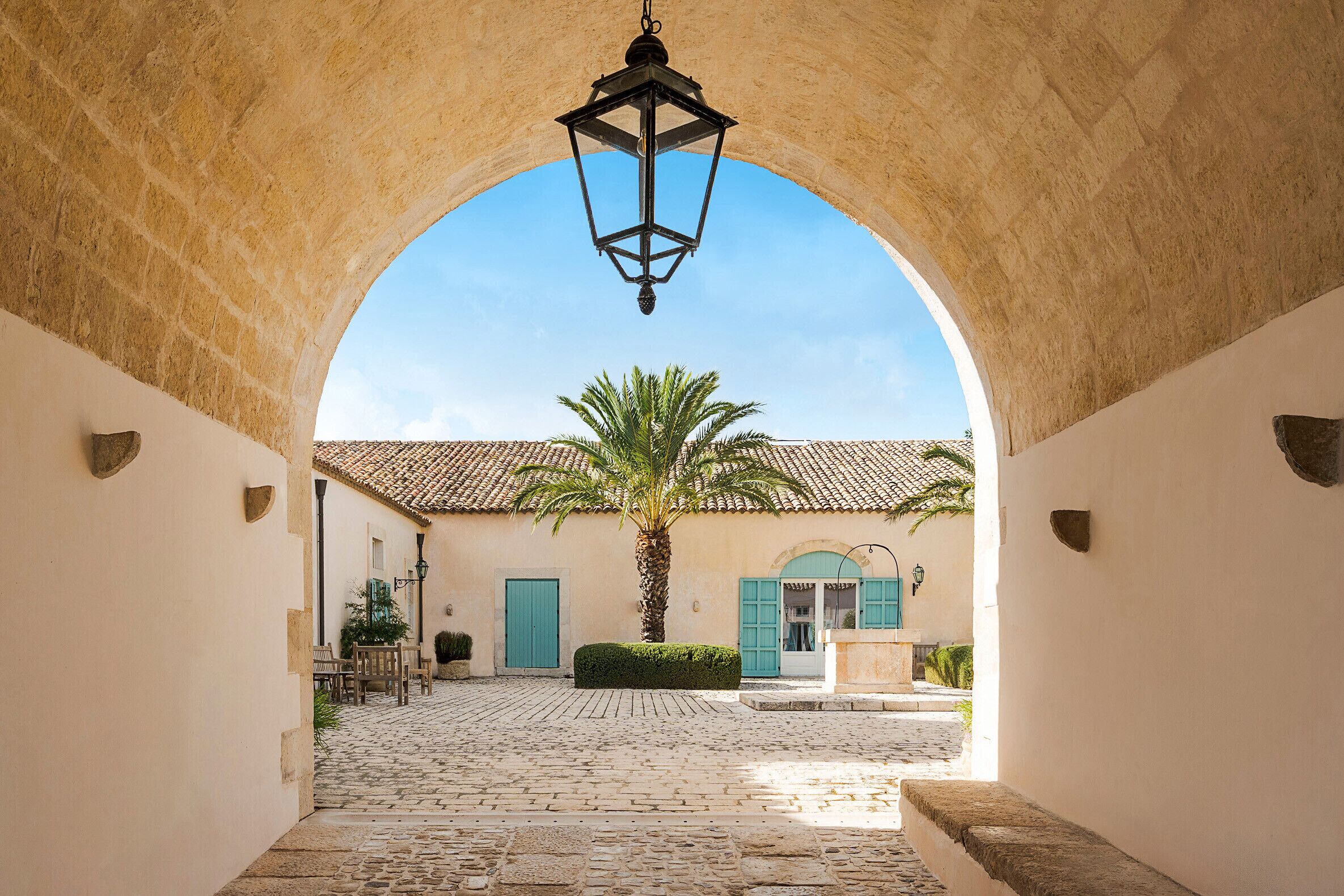 An arched corridor with cobblestone flooring, a palm tree, and a building with a tiled roof and blue shutters.