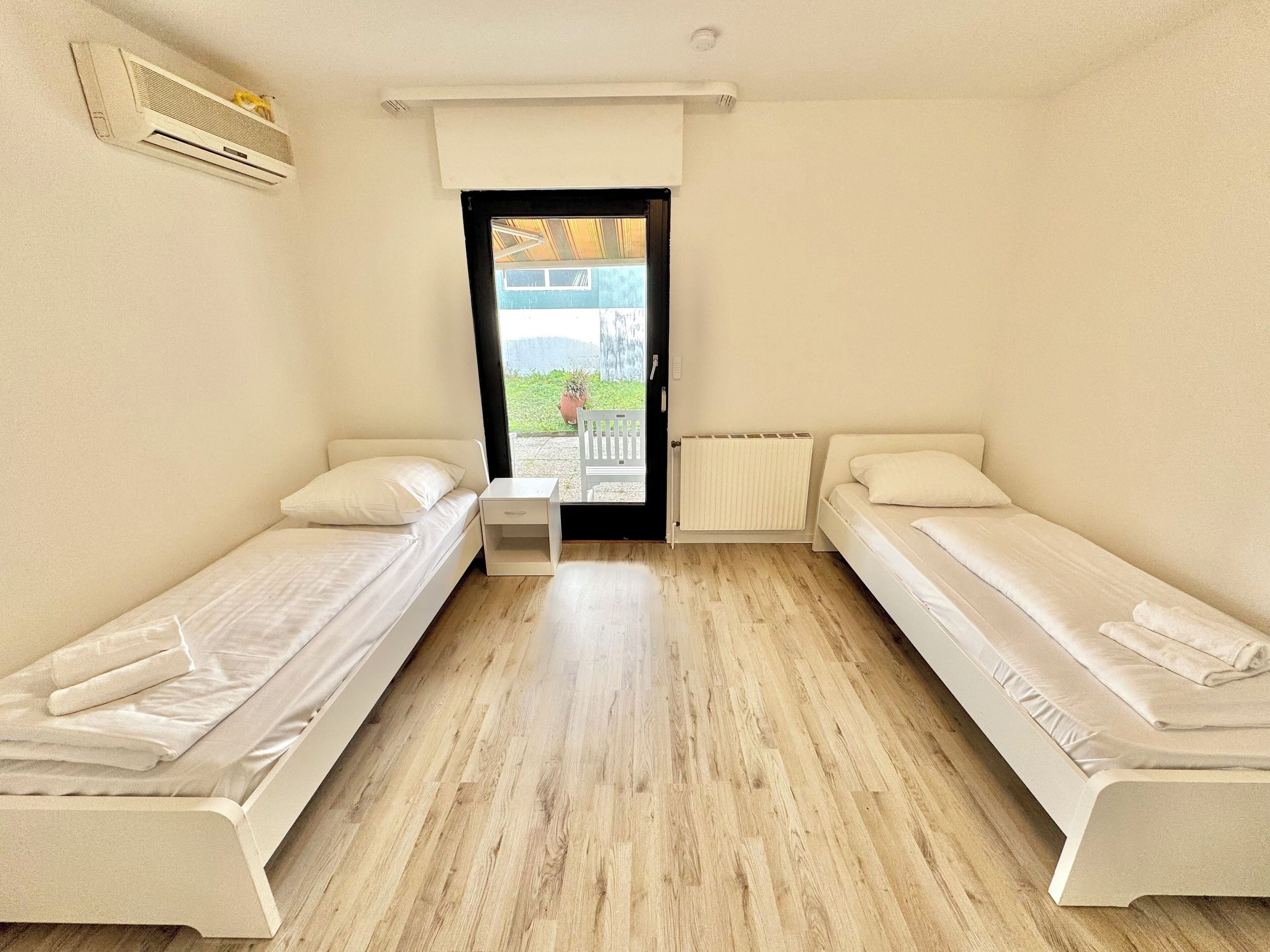 A small, clean room with two single beds, a radiator, and a window with a view of greenery outside.