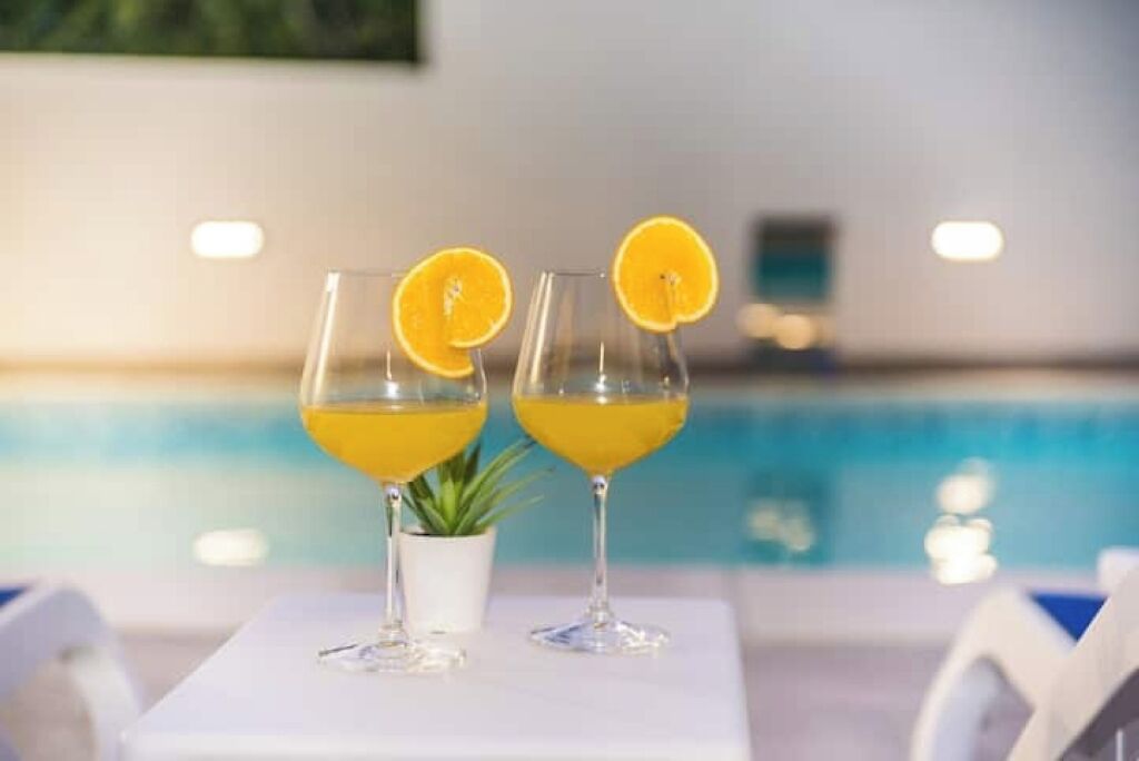 Duas taças de uma bebida amarela com fatias de laranja sobre uma mesa branca perto de uma piscina.