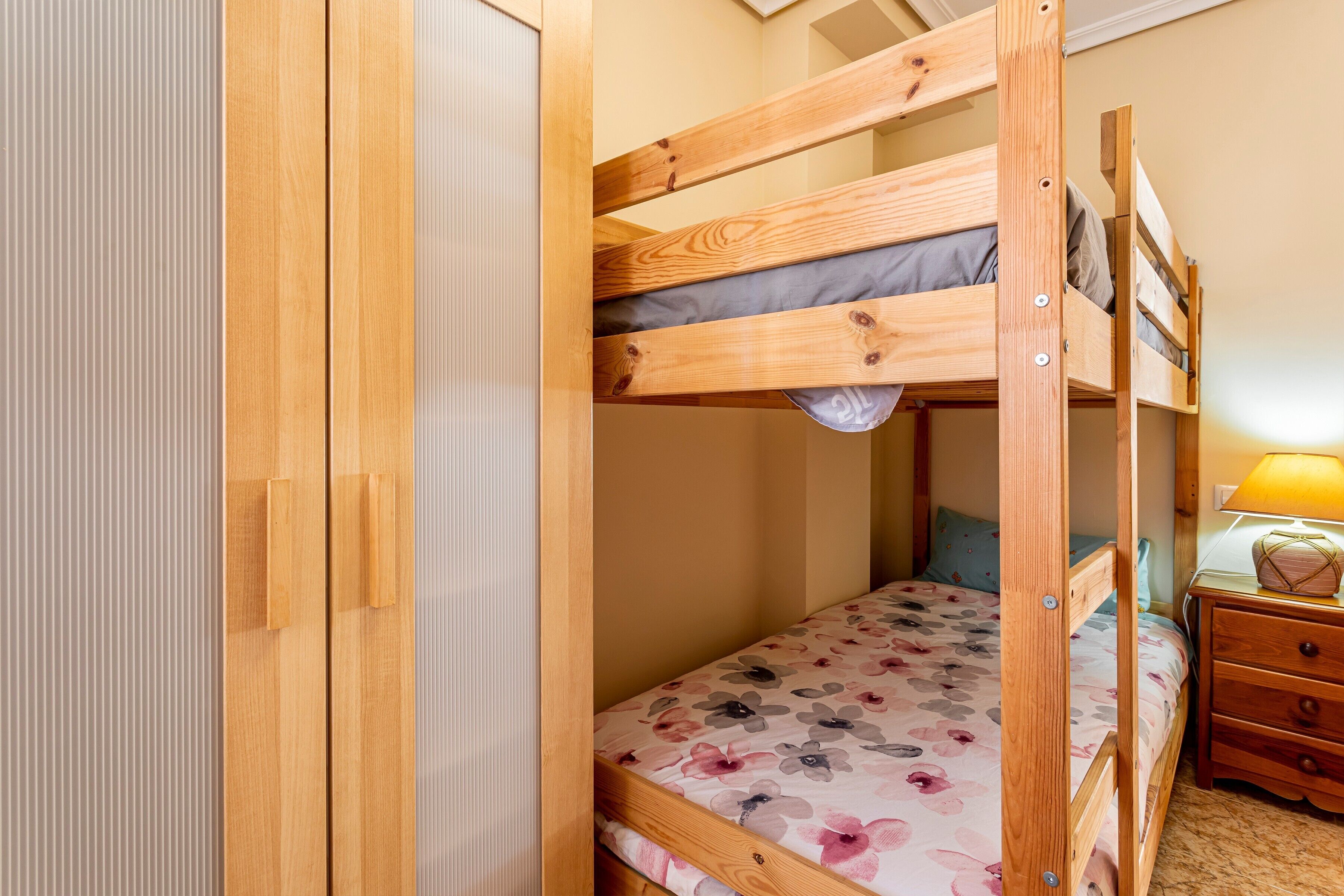Una cama litera con colcha estampada, un armario de madera y una mesita de noche con lámpara.