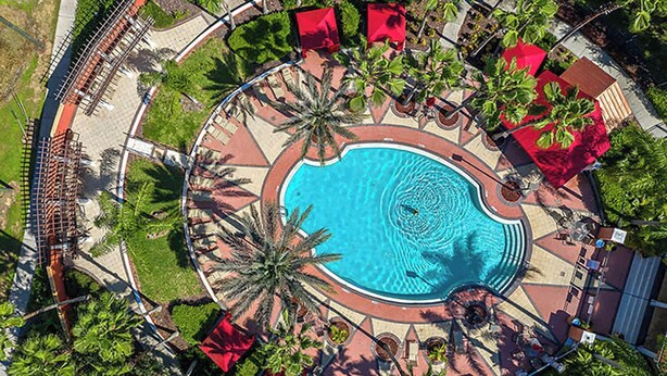 An aerial view of a resort with a central swimming pool, palm trees, and red umbrellas. - Family Resort Home w Pool BBQ Game Room (Davenport)
