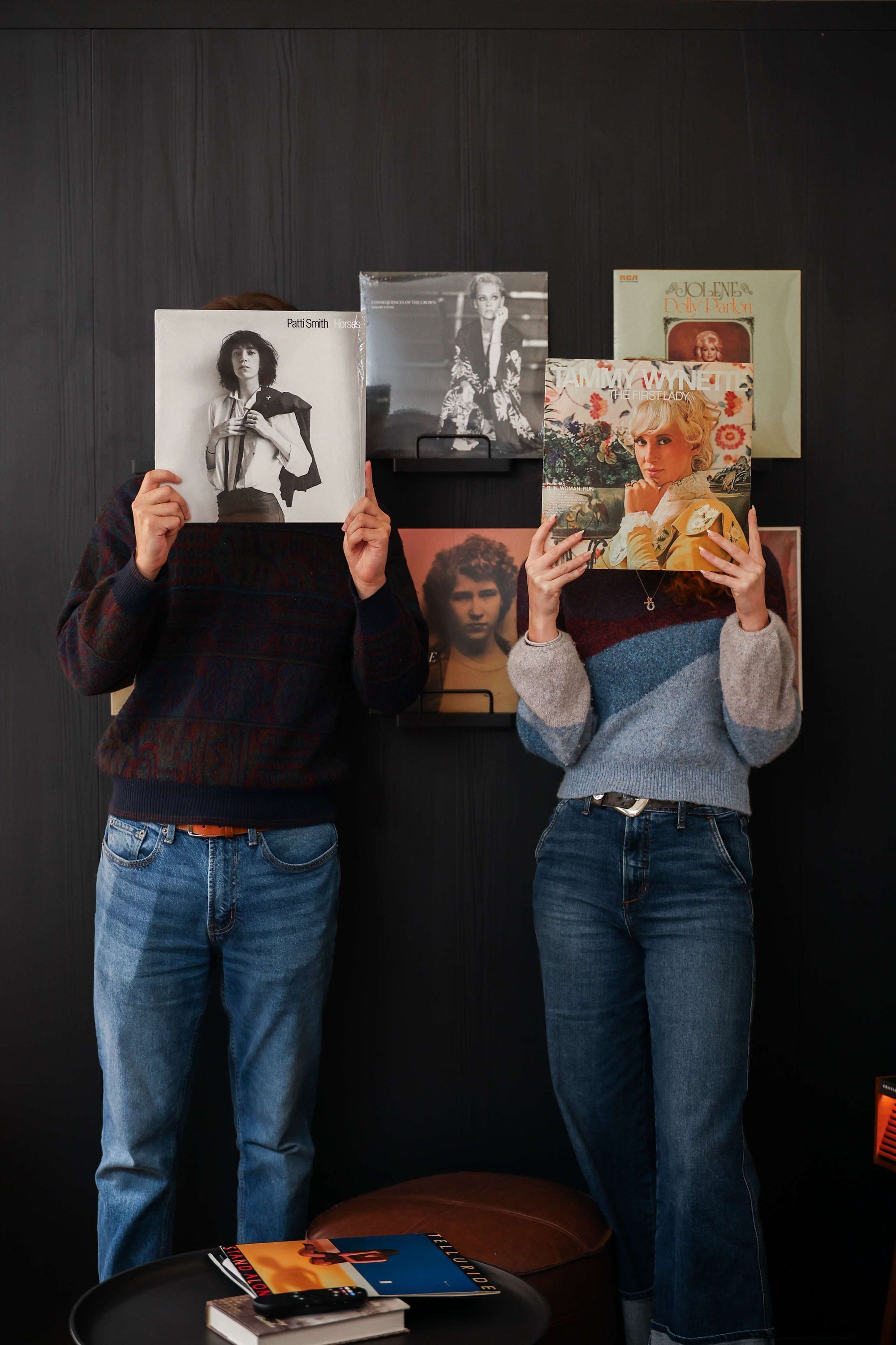 Two individuals holding up records, with a wall-mounted picture frame and a small table with books and a remote control.