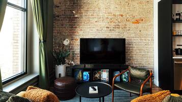 A cozy living room with a brick wall, a flat-screen TV, a leather chair, a small round table, and a window with curtains.