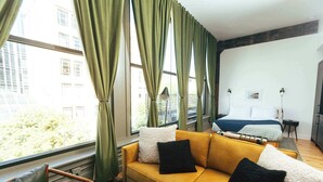 A modern hotel room with a yellow sofa, a bed with a blue blanket, and a view of the city through large windows. - Loft 301 The William - 3rd Ave Views - Carrigans (Birmingham)