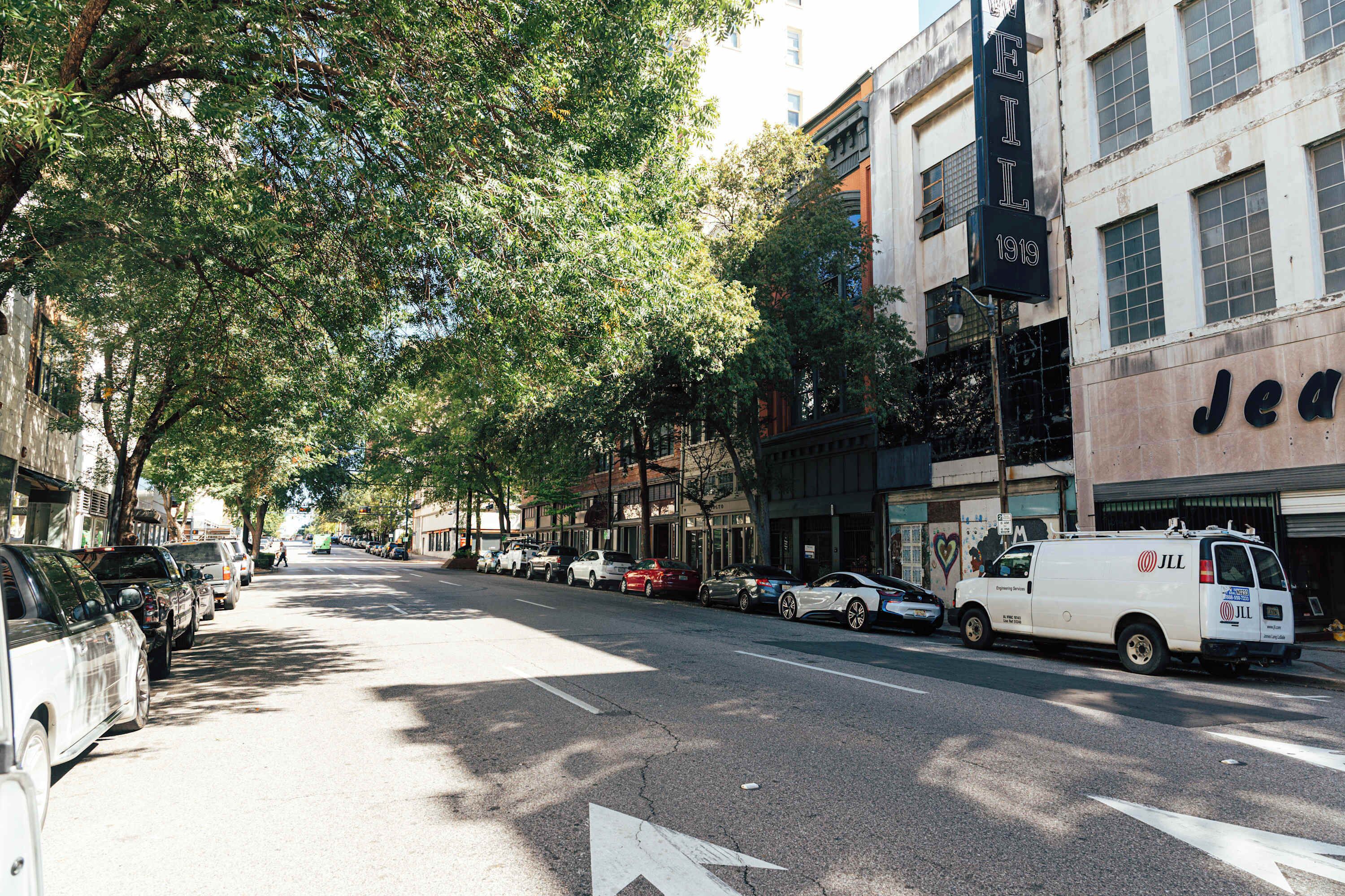 Una calle bordeada de coches aparcados y una camioneta blanca. Se ven árboles y edificios a lo largo de la calle.