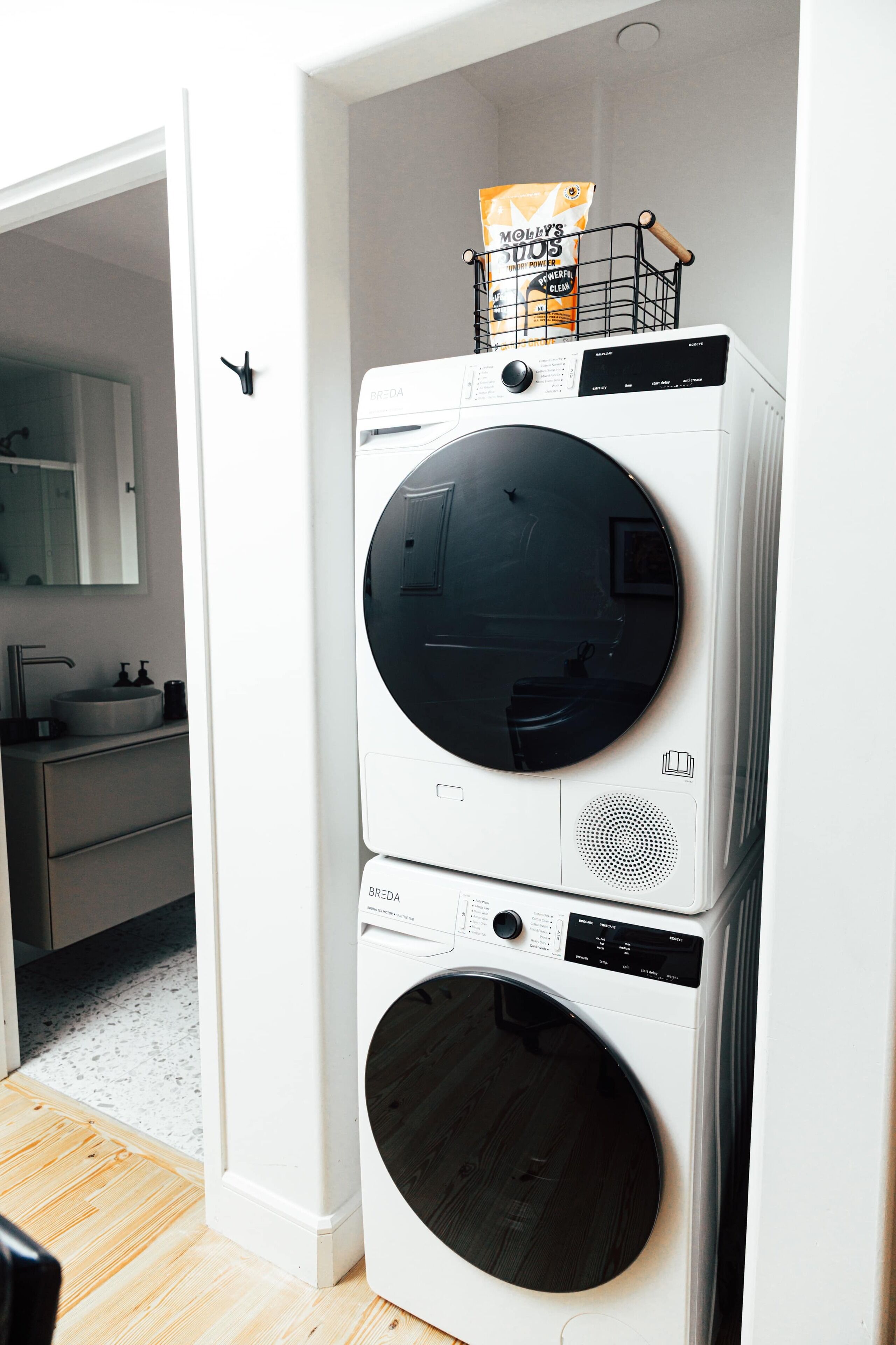 A stacked washer and dryer in a room with a wooden floor, a white wall, and a door leading to a bathroom.