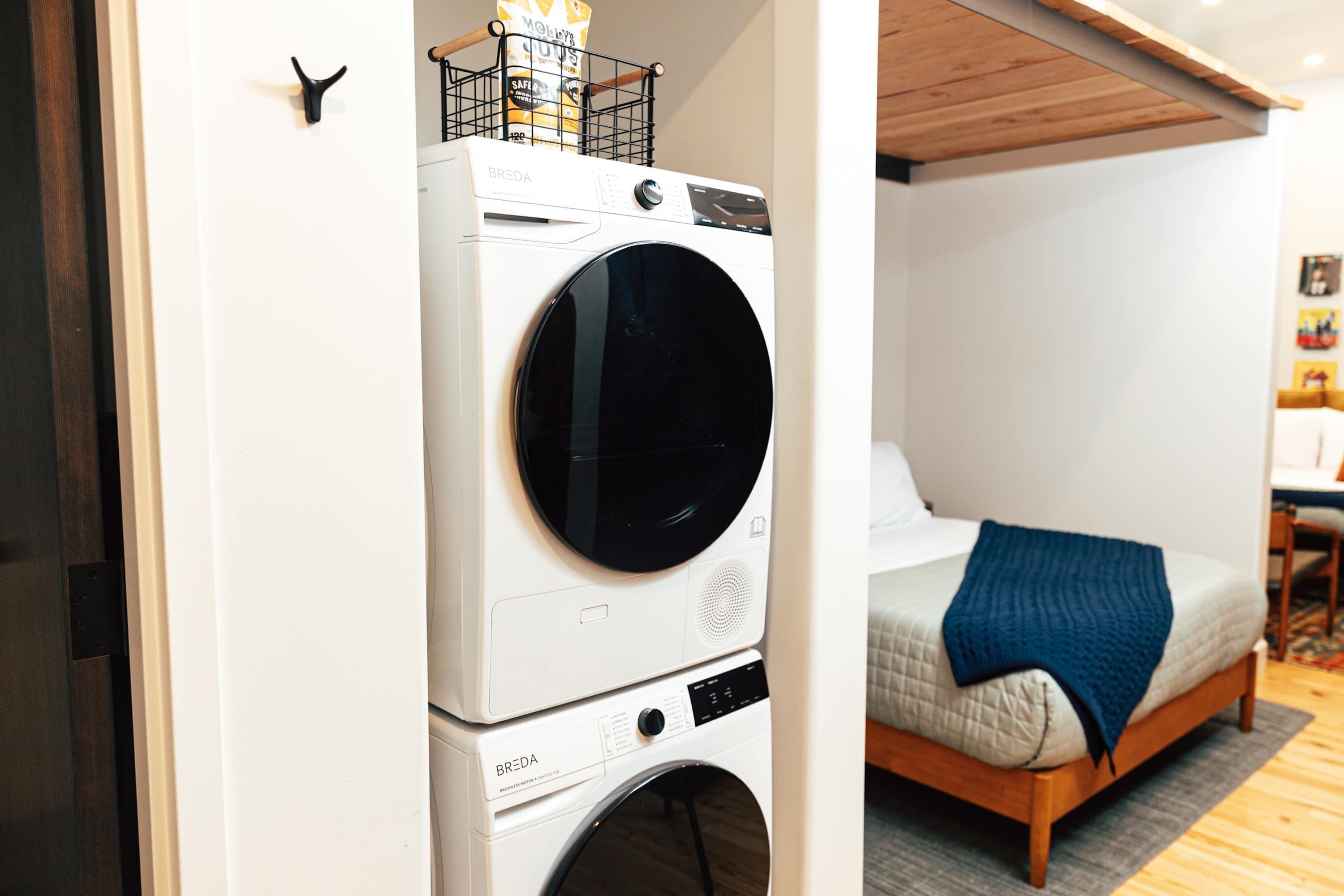 Una habitación pequeña con cama, lavadora y una pequeña mesa con una silla.