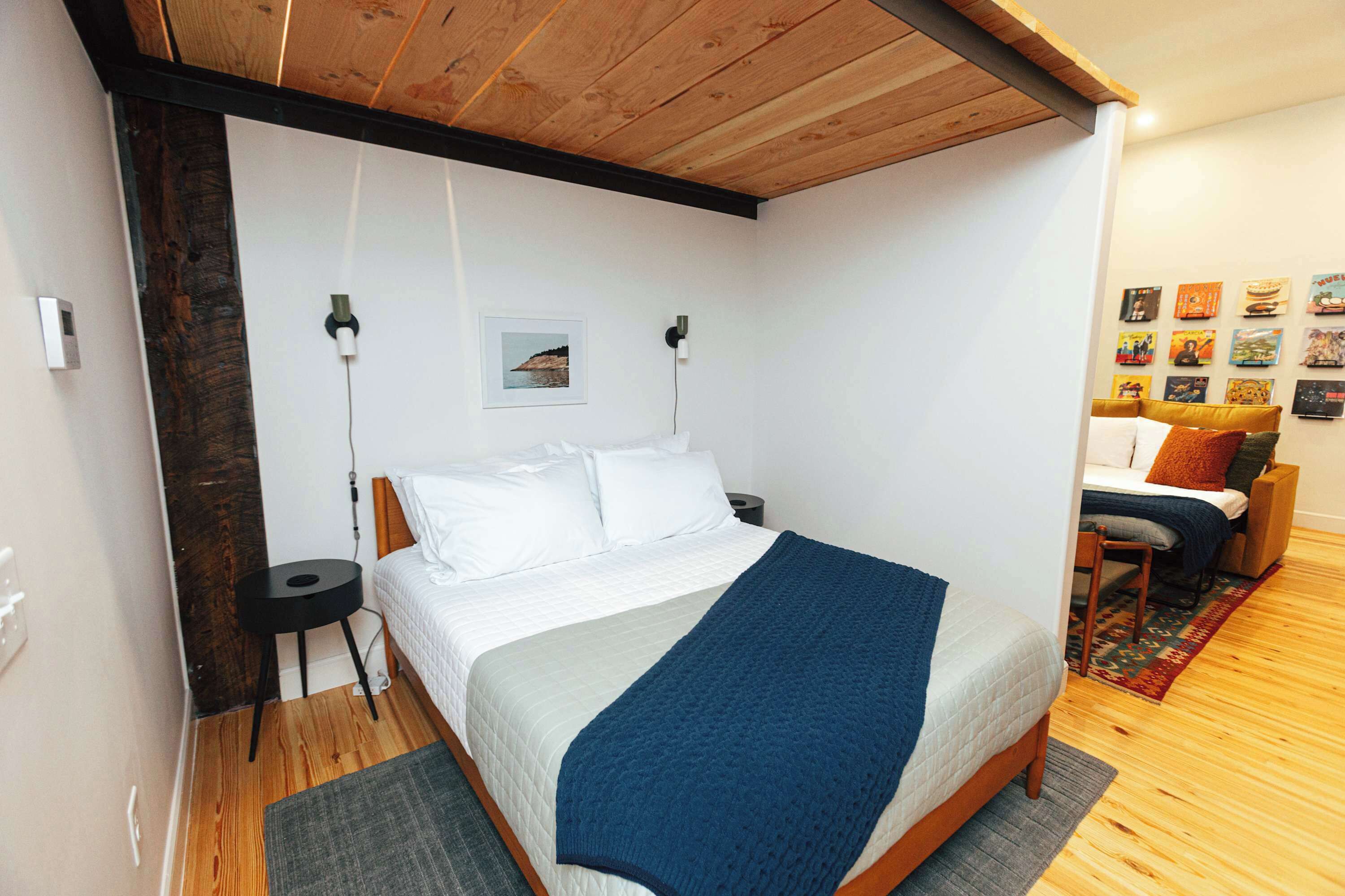 A bedroom with a wooden ceiling, a bed with white bedding and a blue blanket, a small side table, and a wall with framed pictures.