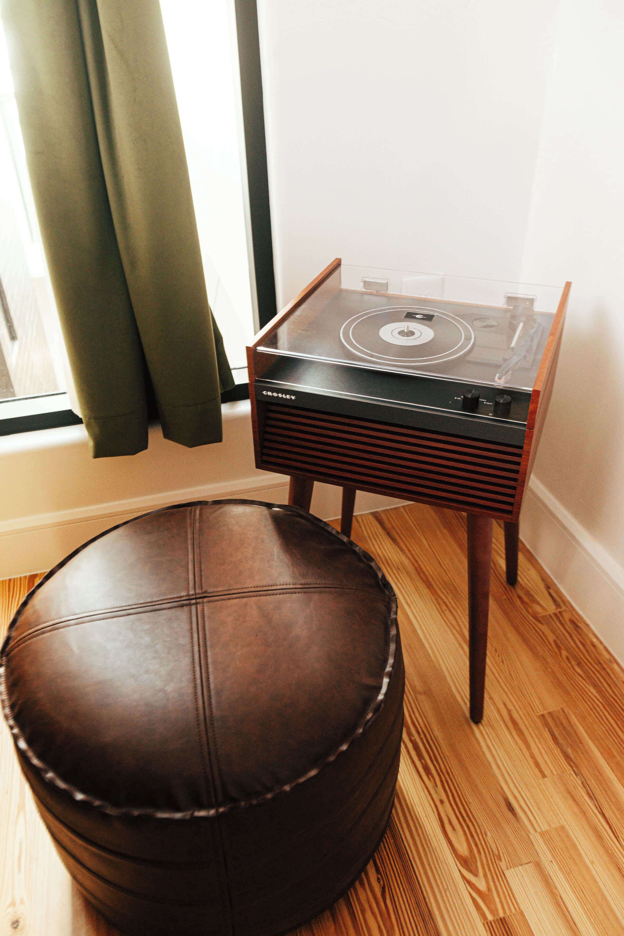 Ein Zimmer mit einem braunen Ledersitzhocker, einem Plattenspieler von Crosley im Vintage-Stil und einem Fenster mit Vorhängen.