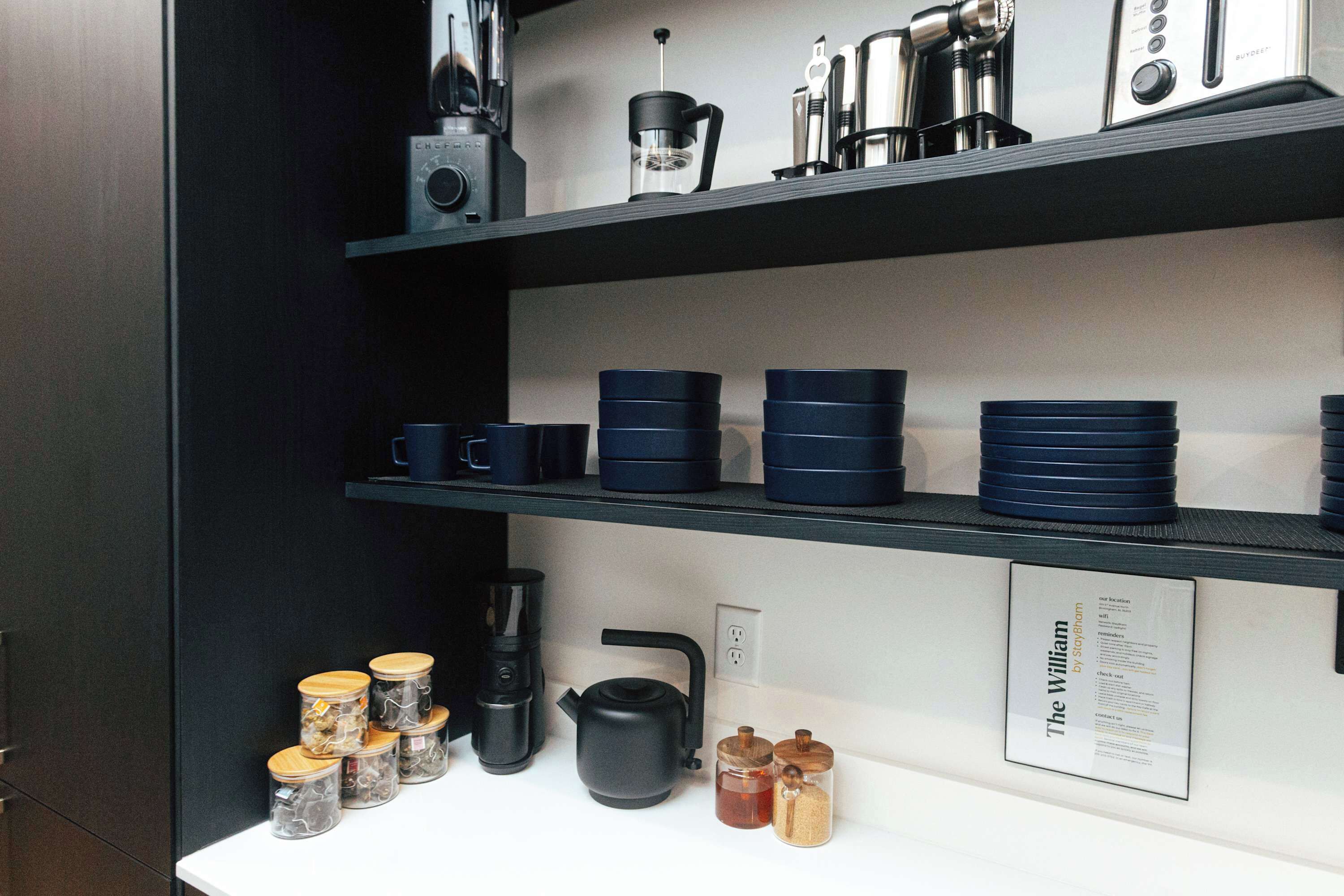 Una cocina con estantes negros que exhiben diversos utensilios de cocina, incluyendo una tetera, una cafetera y recipientes de almacenamiento.