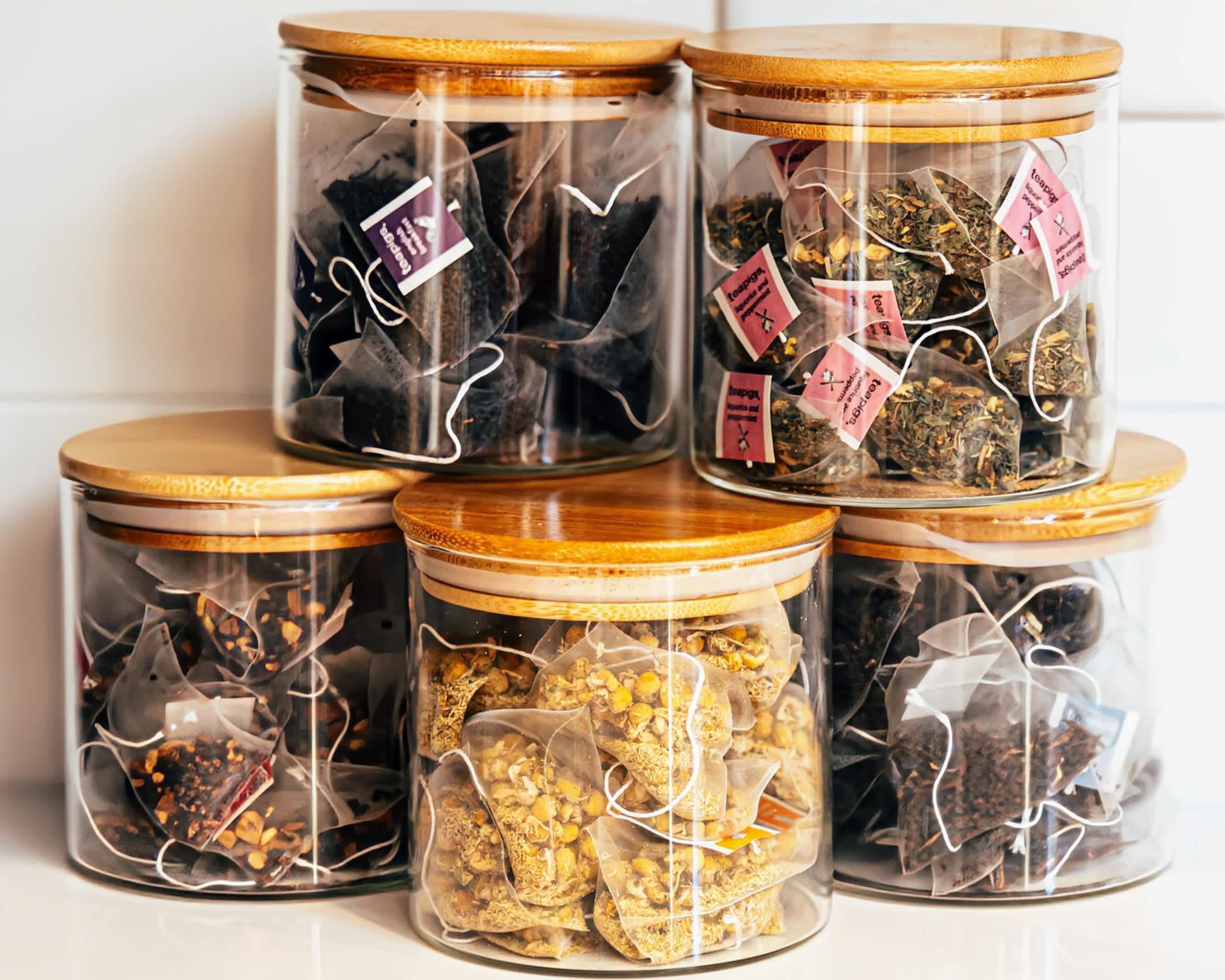 Cinco frascos de vidrio con tapas de madera, cada uno con diferentes tipos de hojas de té, dispuestos en una estantería.
