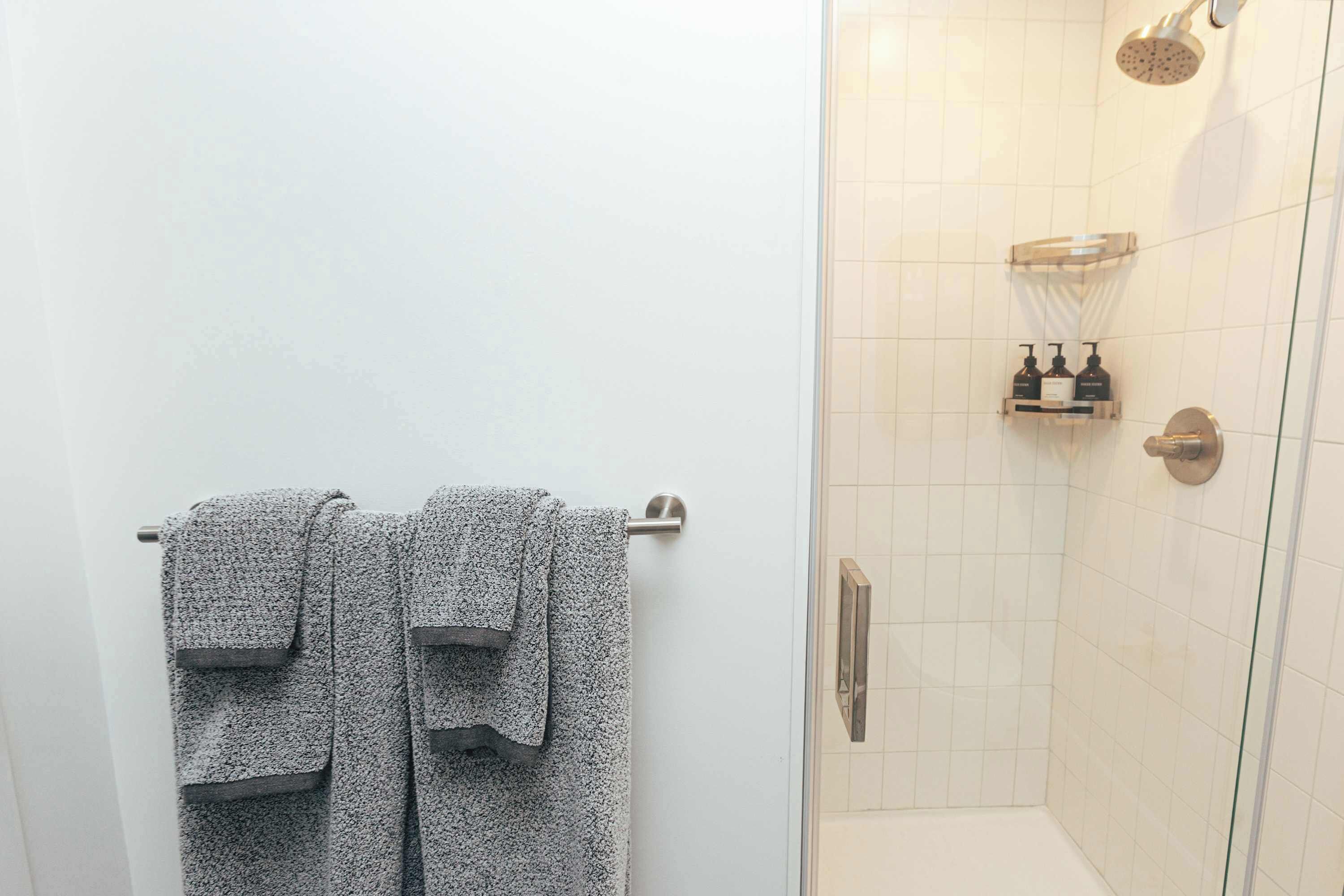 Un baño con dos toallas colgadas en un perchero, una ducha con puerta de vidrio y un estante con artículos de aseo.