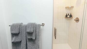 Un baño con dos toallas colgadas en un perchero, una ducha con puerta de vidrio y un estante con artículos de aseo.