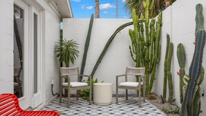 Ein kleiner Patio-Bereich mit zwei Holzstühlen, einem runden Tisch und verschiedenen Kakteen vor einer weißen Wand.