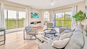 A spacious living area with a large sofa, a coffee table, and a TV mounted on the wall. There are large windows offering a view of the outdoors. - Upstay Condo w Pool Sauna Sleeps 8 (Winter Garden)