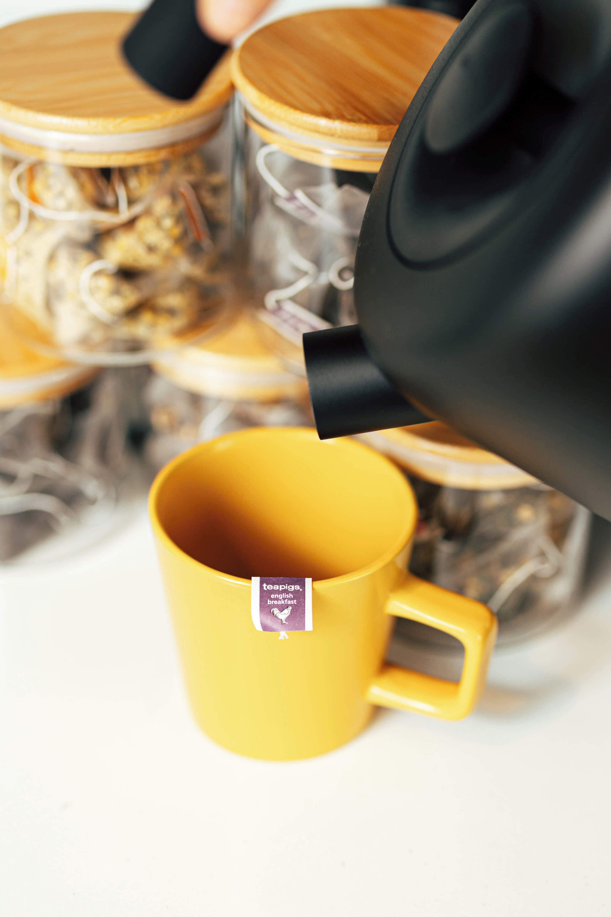 Una taza amarilla con etiqueta de bolsita de té, una tetera negra vertiendo en ella, y un tarro de madera con tapa al fondo.