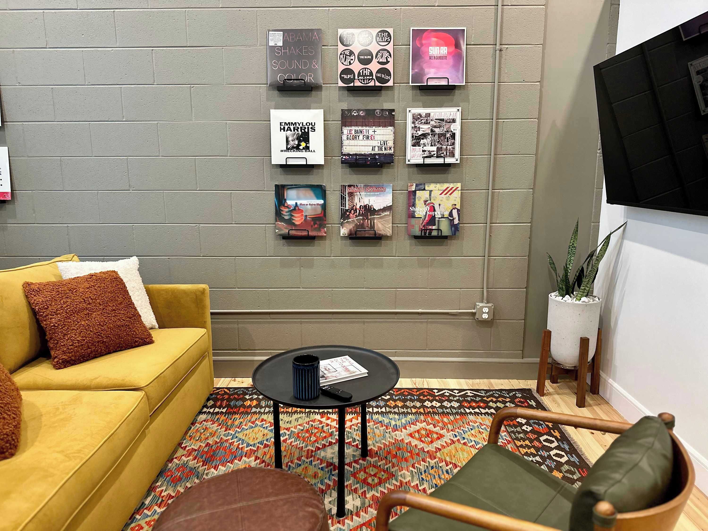 Una habitación con sofá amarillo, una mesita redonda pequeña, un tapete estampado y una pared decorada con carteles enmarcados.