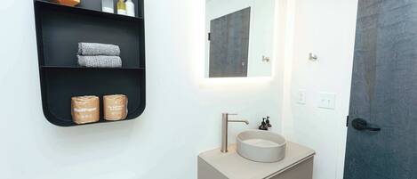 Un baño moderno con un lavabo blanco, un espejo y una estantería negra con artículos de aseo.