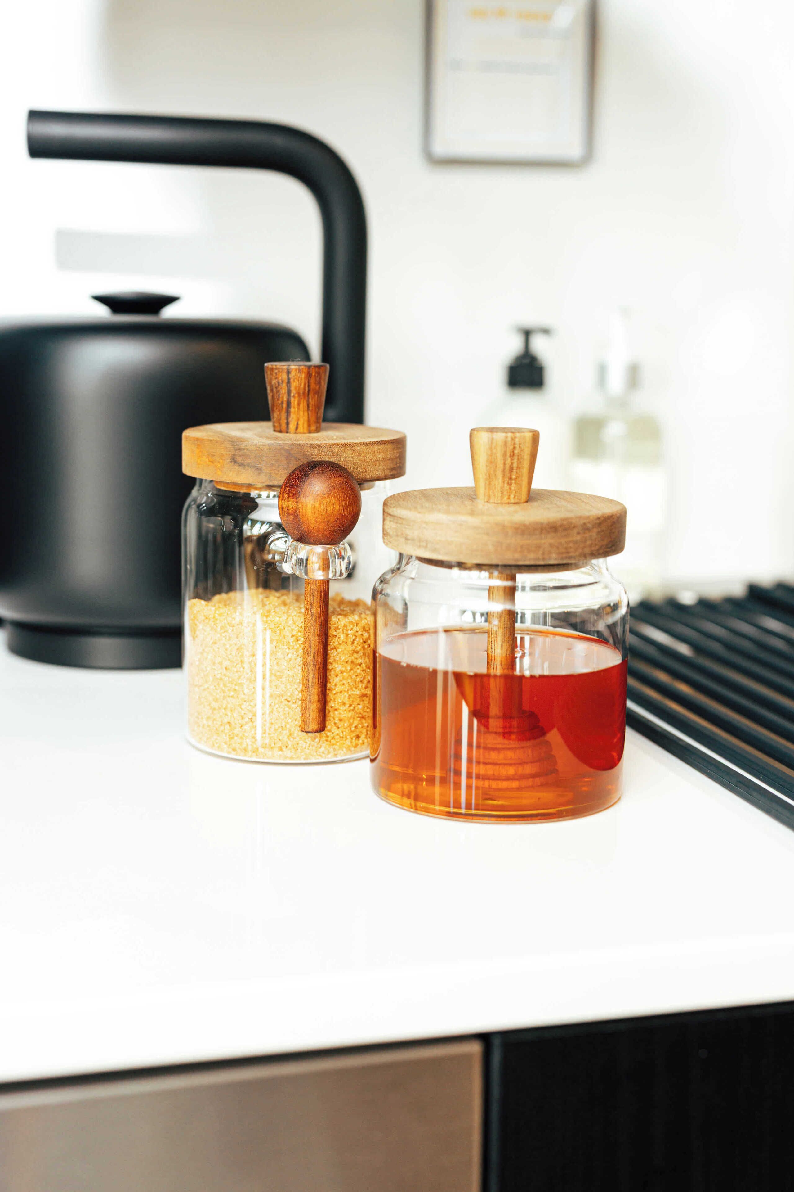 Two glass jars with wooden lids on a white countertop, one containing grains and the other honey, with a black kettle in the background.