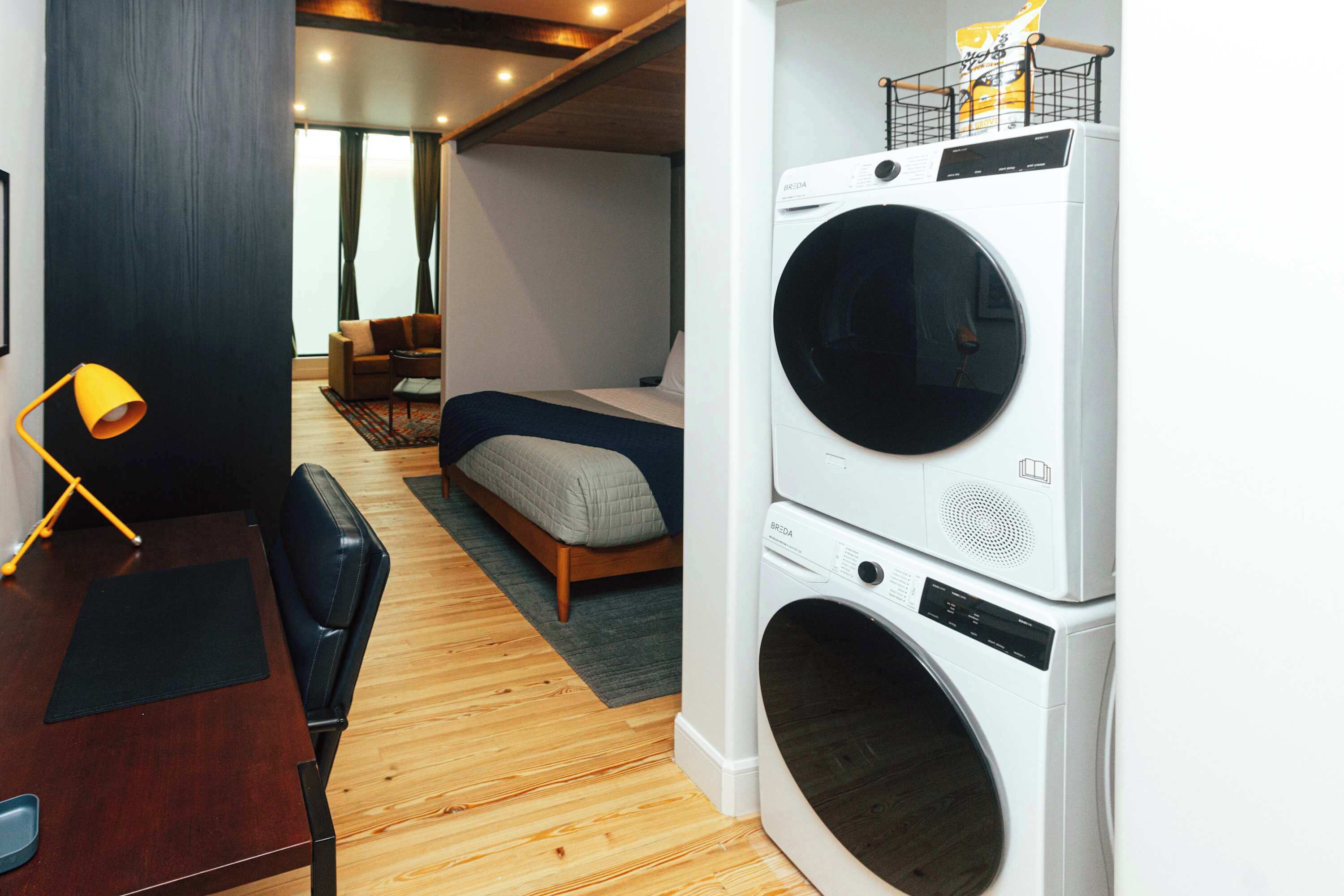 A compact hotel room with a bed, a desk, a chair, a washing machine, and a view through a window.