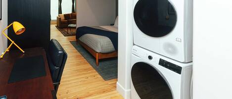 A compact hotel room with a bed, a desk, a chair, a washing machine, and a view through a window.