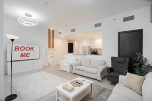 A modern living room with a sofa, armchairs, a coffee table, and a TV displaying "BOOK ME". - Stayshort - Serene 2-bedroom With Relaxing Balcony Atmosphere in Liwan (Dubai)
