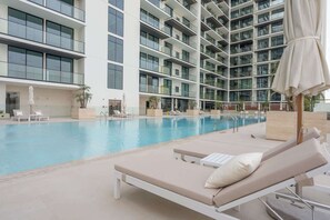 A modern hotel with a large indoor swimming pool, multiple lounge areas with sunbeds, and a high-rise building with balconies. - Stayshort - Sophisticated JVC Apartment Sleeps 4 With Balcony (Dubai)