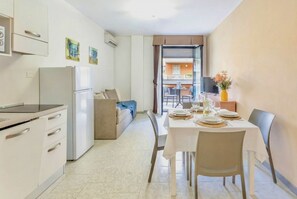 Una cocina moderna con refrigerador, estufa y gabinetes. Hay una zona de comedor con una mesa preparada para dos personas, y un sofá al fondo.
