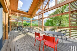 Eine Holzterrasse mit Tischen und Stühlen, großen Fenstern und Blick auf grüne Bäume und Berge.