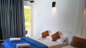 Habitación de hotel con dos camas, cada una con una colcha azul y blanca, y cabeceros de madera. Se ve una ventana con cortinas, una lámpara de pared y una pequeña planta en maceta junto a la ventana.